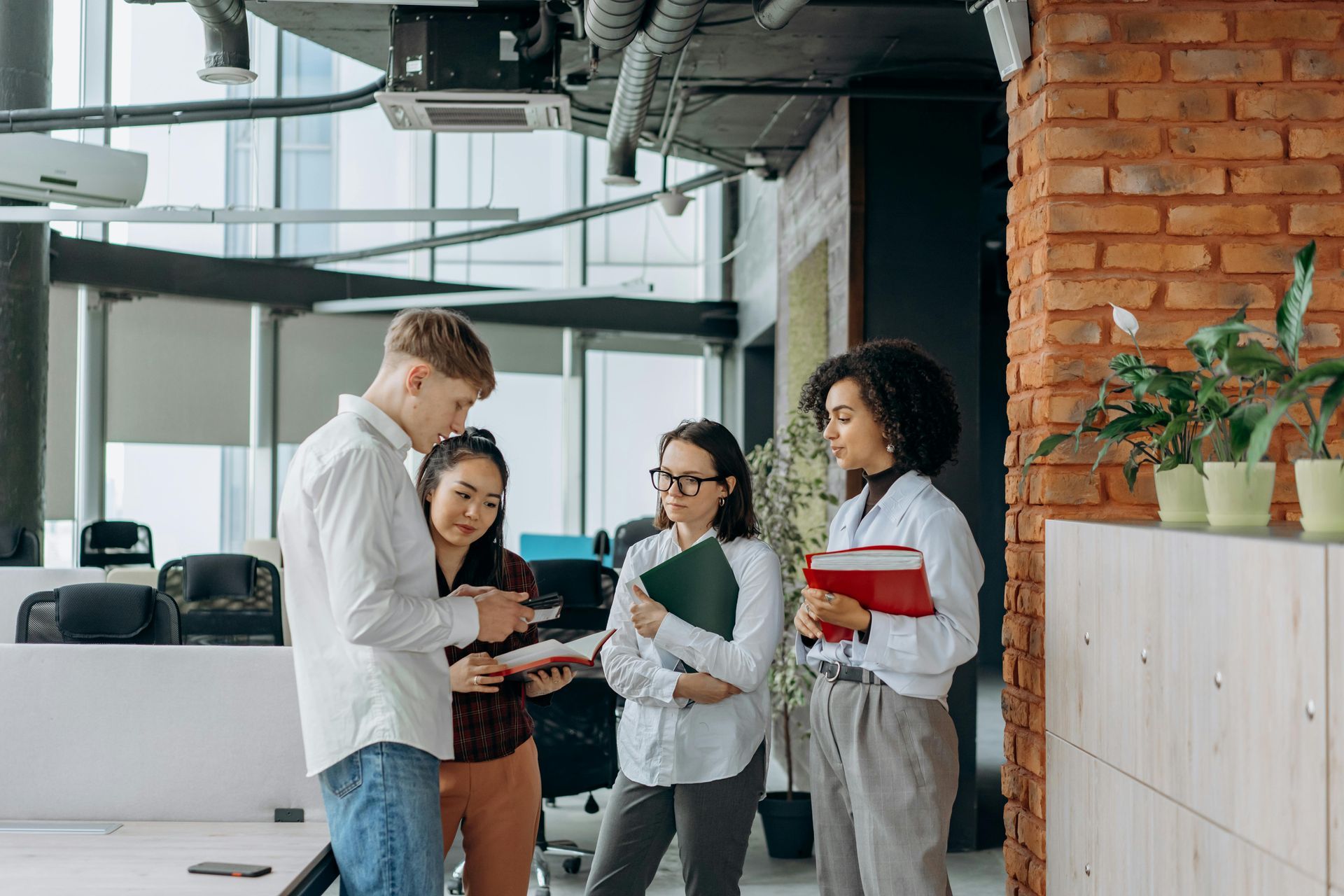 Four coworkers discussing notes in a modern office with brick wall and glass stairs