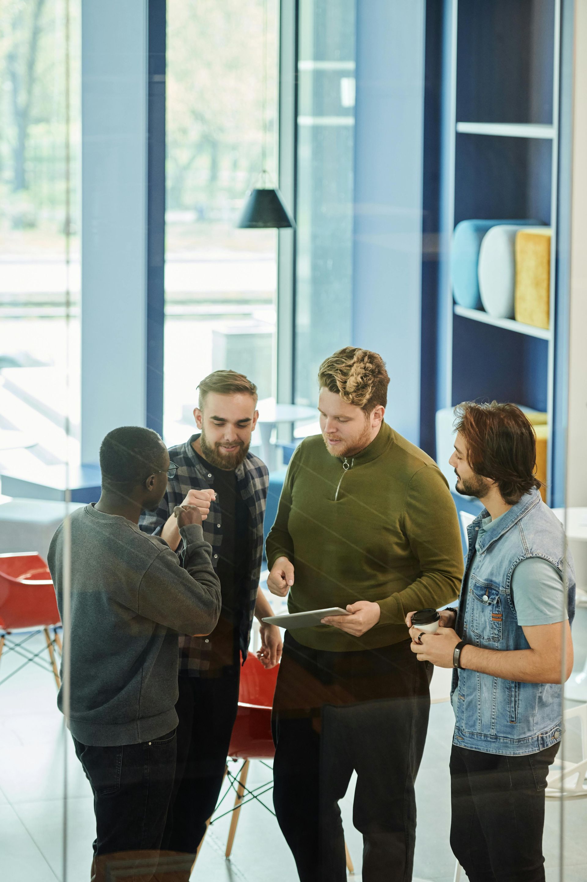 Four coworkers gather in a modern office, discussing papers and a tablet near blue shelves.