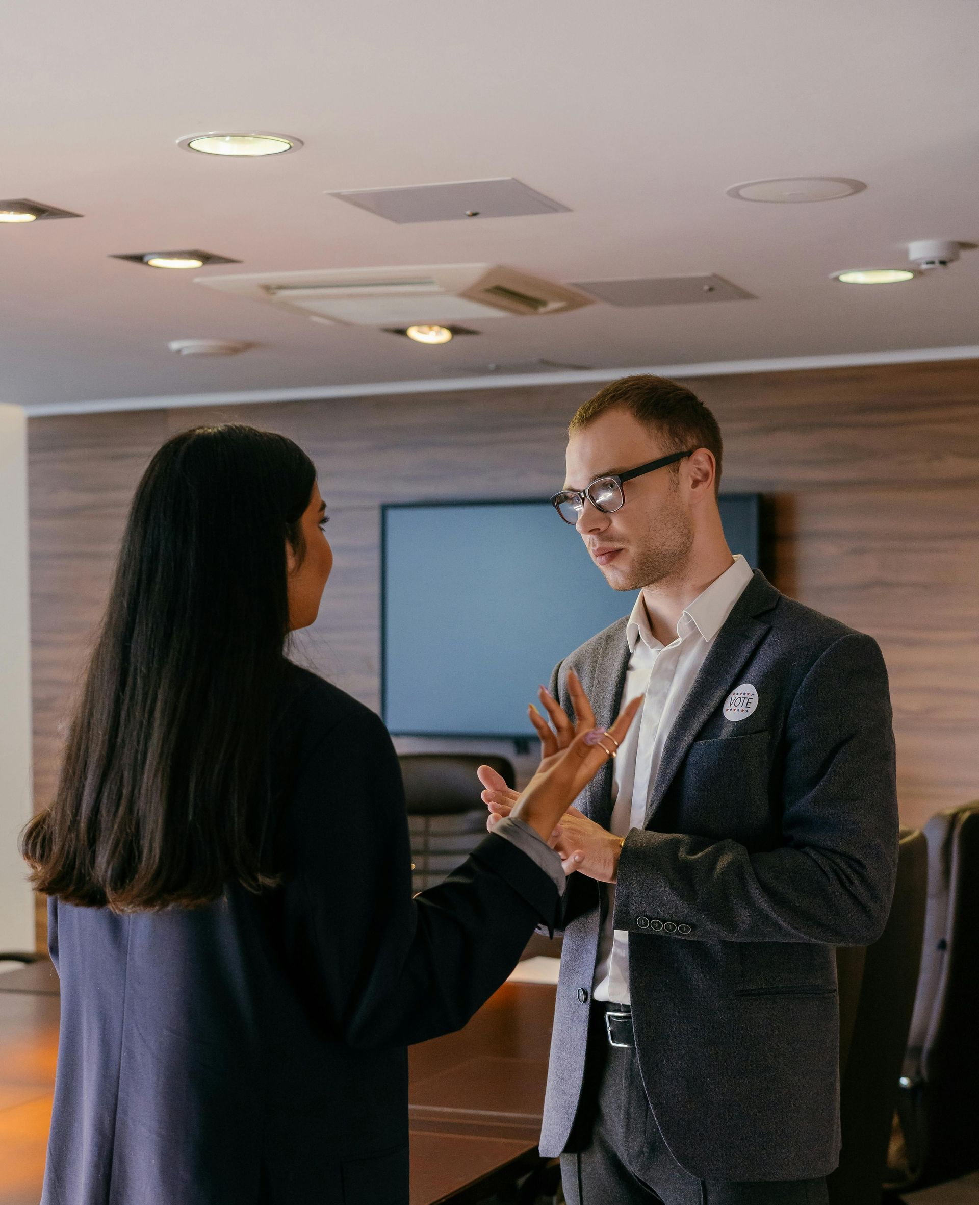 Two colleagues chatting in a modern office, gesturing during a discussion near a conference room screen.