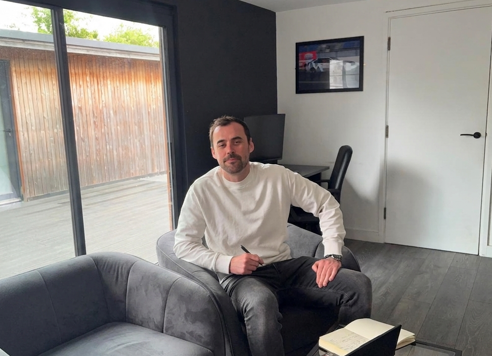 Man seated on a gray sofa in a bright modern living room with large windows and a white door.