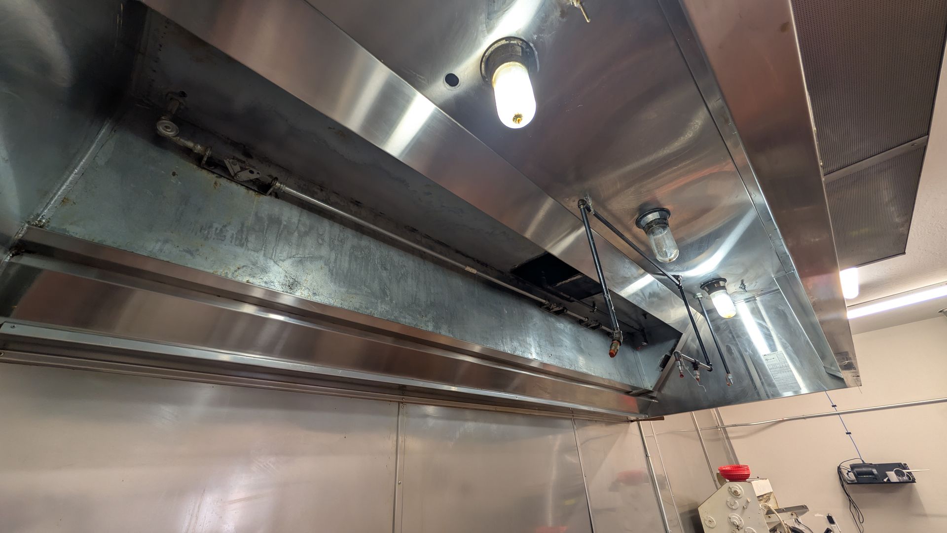 Stainless steel commercial kitchen exhaust hood with lights and vents above a tiled wall.