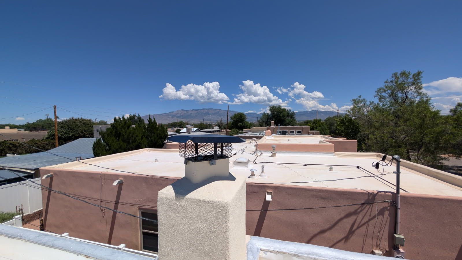 View of a flat rooftop with a chimney and blue sky with clouds. Distant mountains are visible in the background.