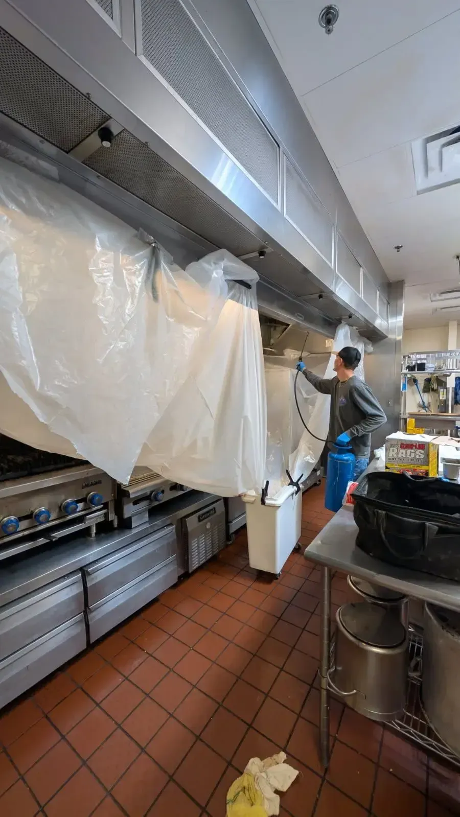 A person sprays a commercial kitchen exhaust hood, contained within plastic sheeting. The kitchen has stainless steel appliances and red tile.