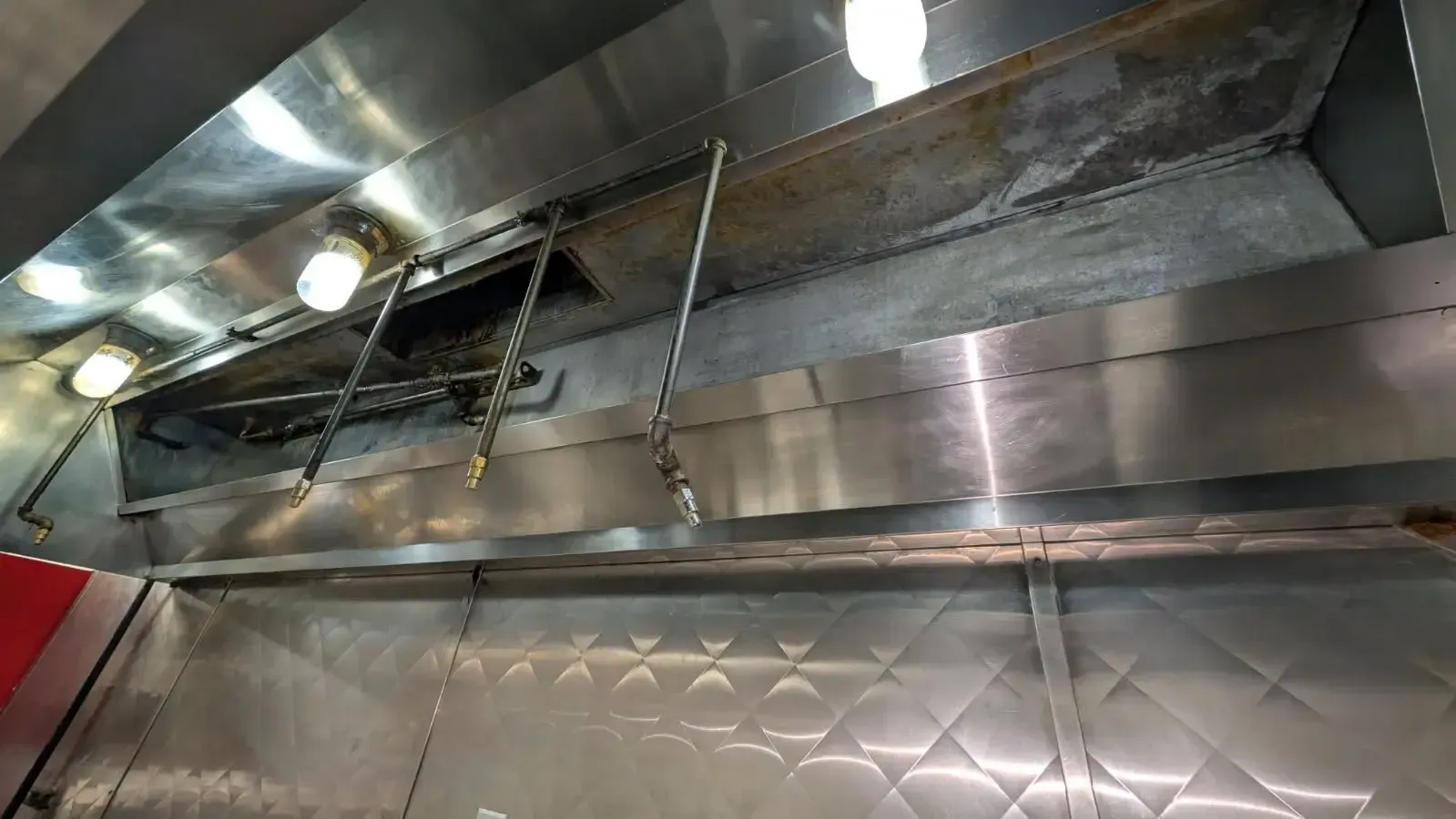 Interior of a stainless steel commercial kitchen hood. It's dirty, with grease buildup visible. There are lights and a vent.