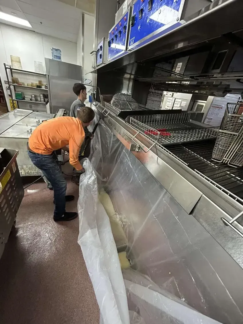 Two people cleaning a commercial kitchen vent, one in an orange shirt. The setting is a kitchen with stainless steel equipment.