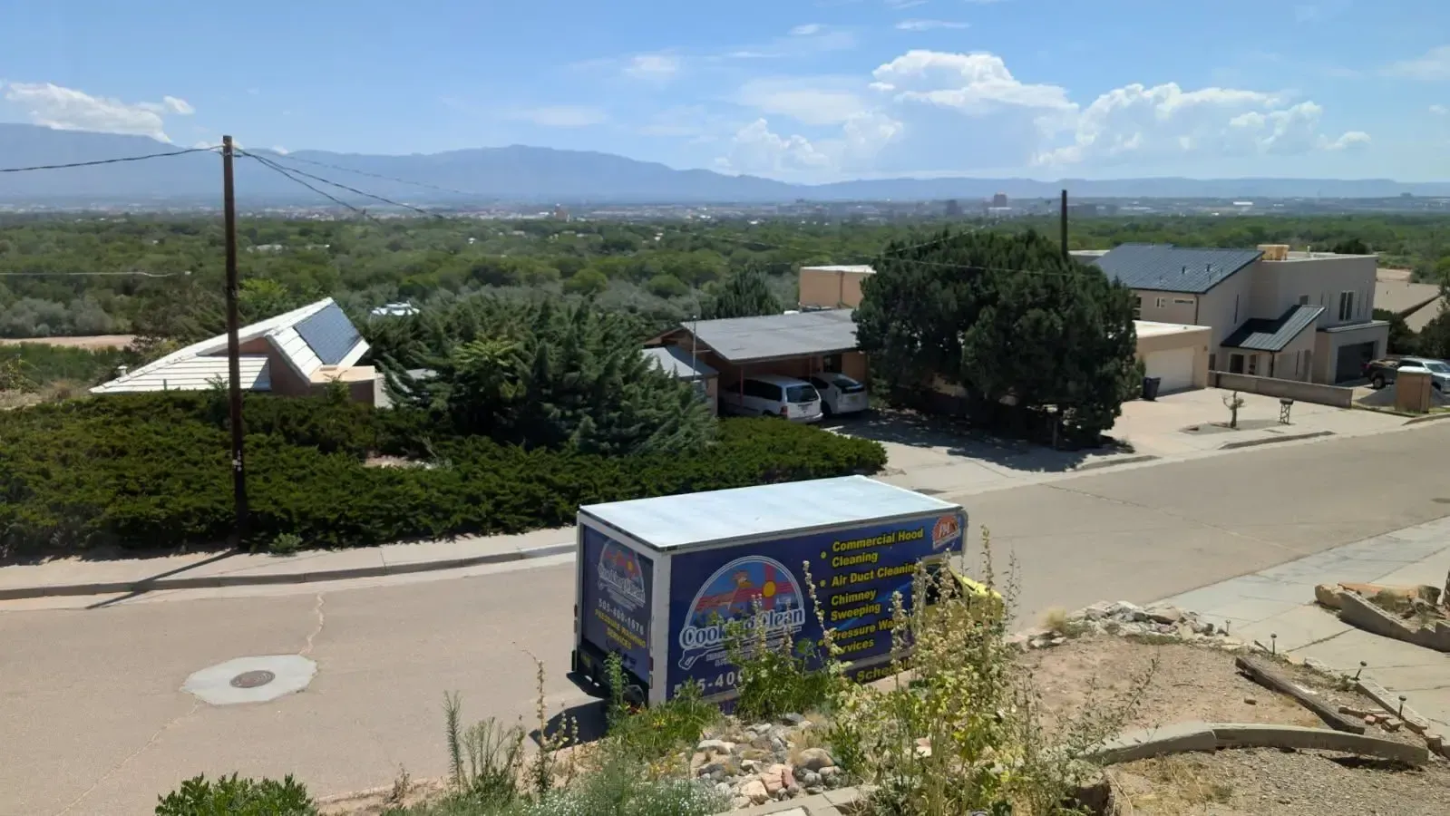 A moving truck parked on a residential street with houses, trees, and mountains in the background under a sunny blue sky.