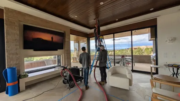 Two people on a tall ladder working on the ceiling of a living room with large windows and a scenic view.