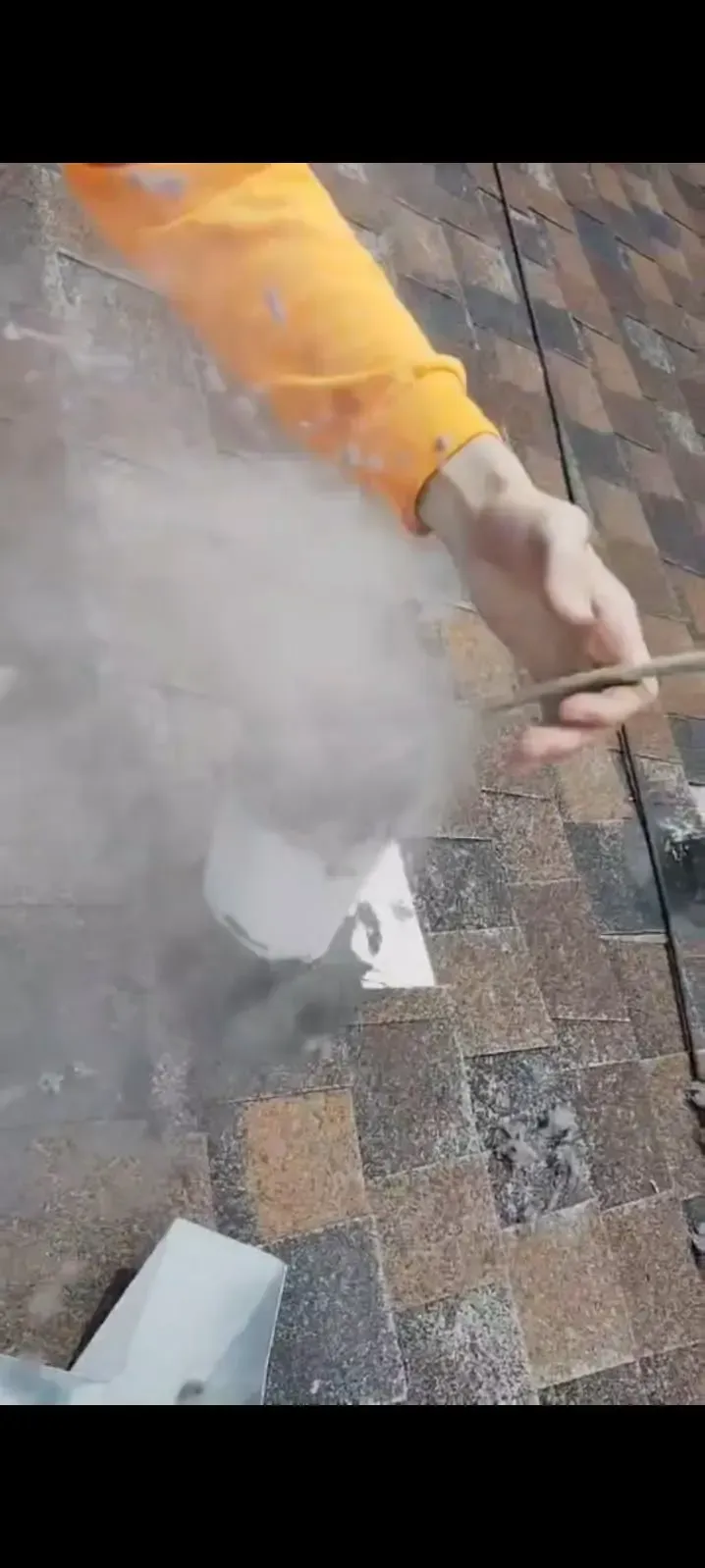 Person in orange shirt working on a roof, using a tool that emits a cloud of smoke or vapor.