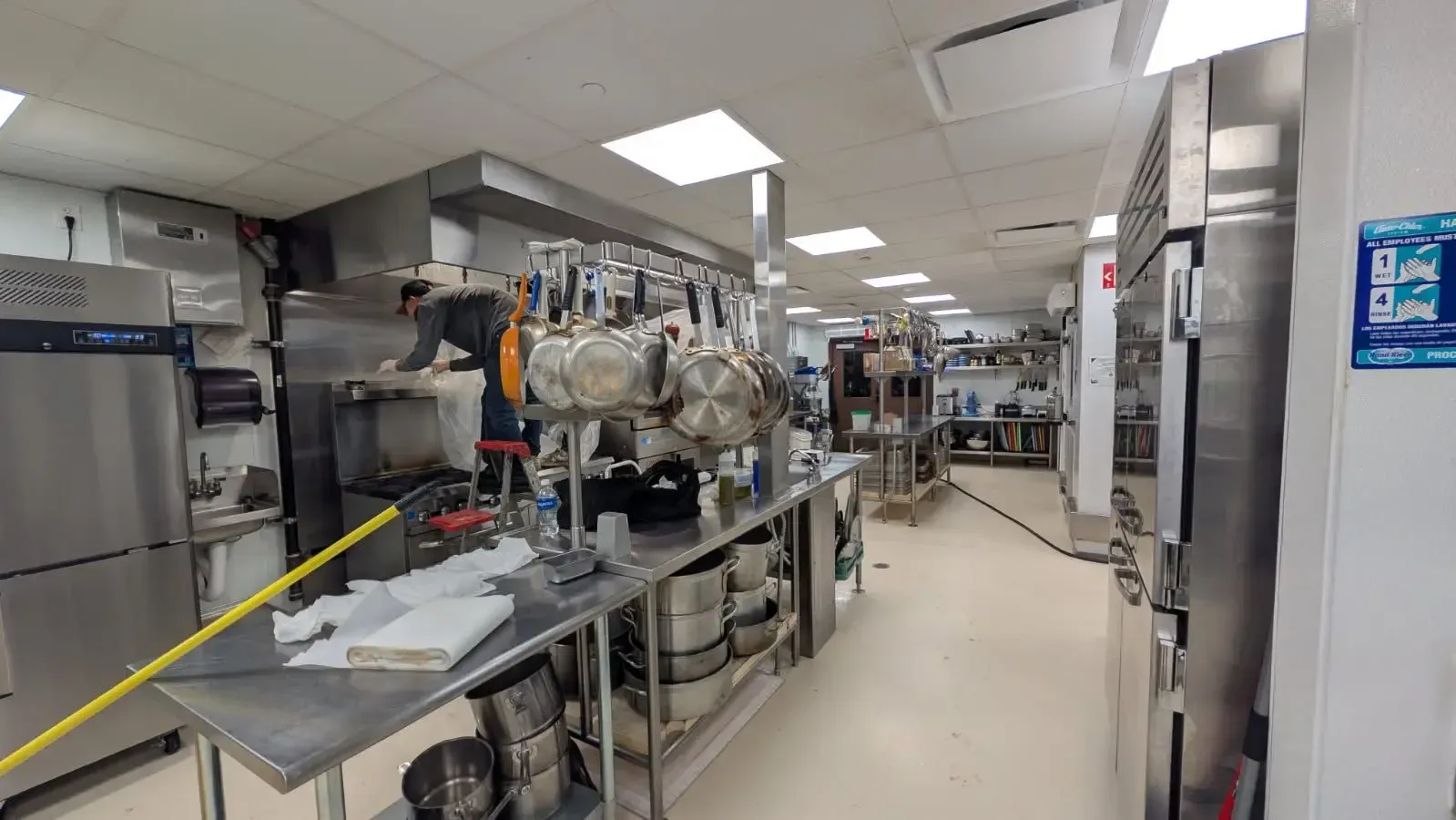 A commercial kitchen with stainless steel appliances, prep tables, and hanging pots. Bright overhead lights illuminate the long, narrow space.