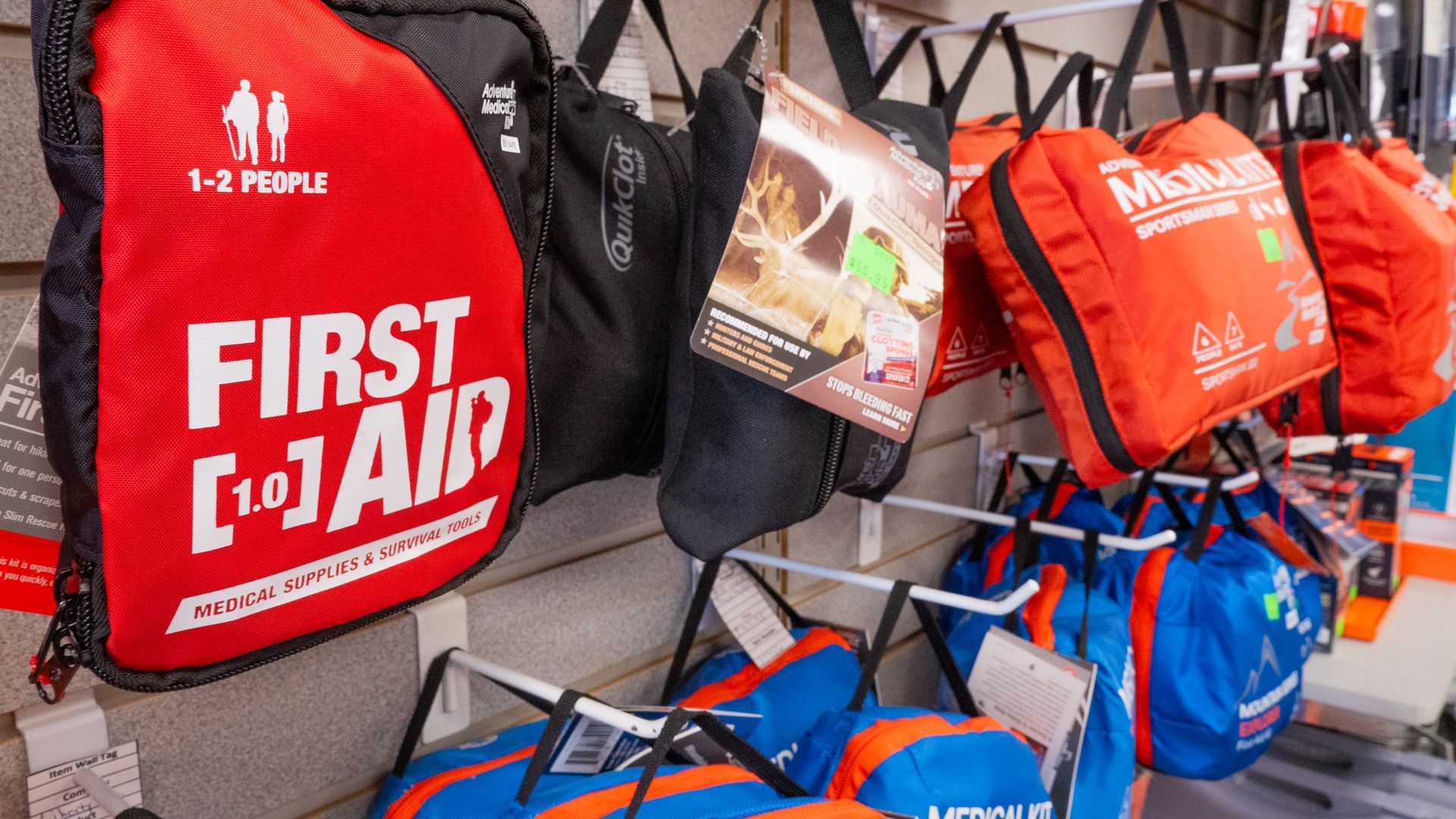 First aid kits for 1-2 people and other supplies are displayed on shelves in a store, with red and blue bags prominent.