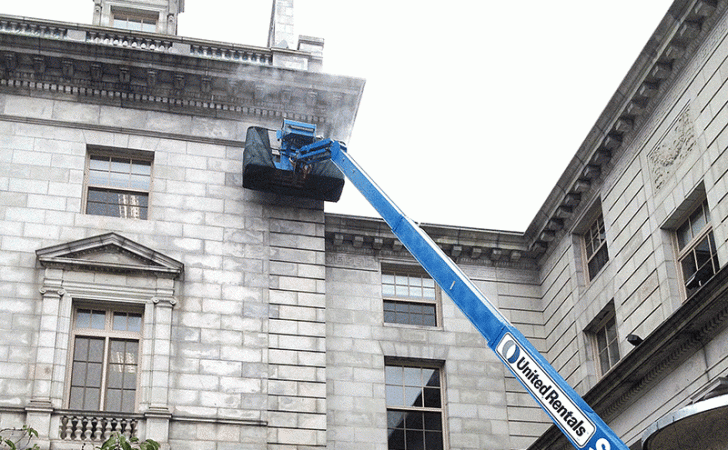 A blue United Rentals boom lift reaches toward the corner of a multi-story stone building to perform maintenance work.