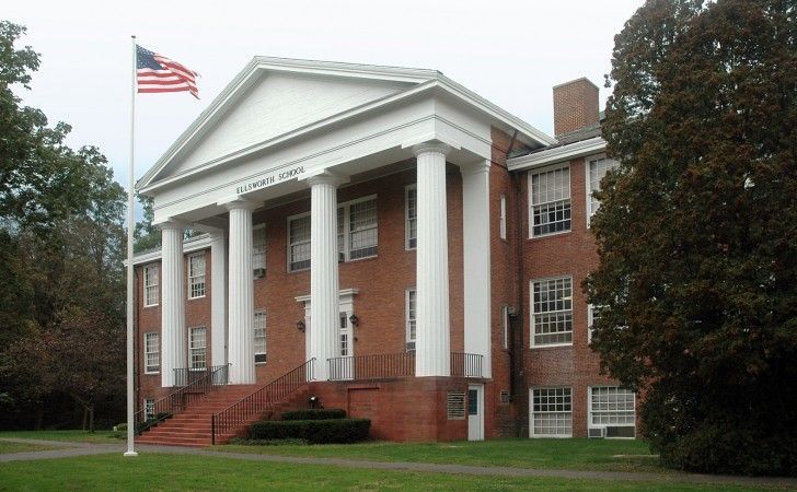 A brick building with a classical white portico, columns, and a triangular pediment, featuring a flag on a pole nearby.
