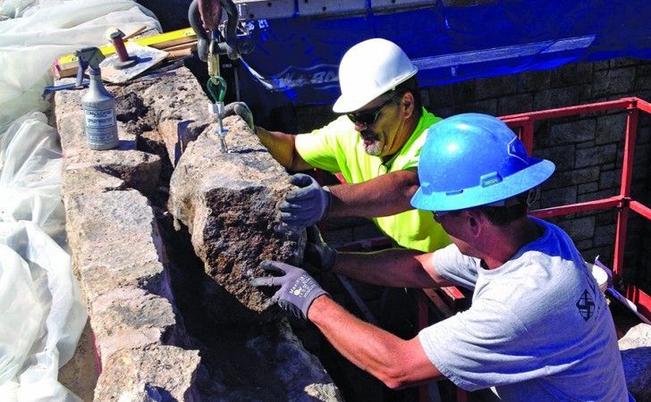 Two construction workers wearing hard hats use a crane to position a large, textured stone block onto a masonry wall.