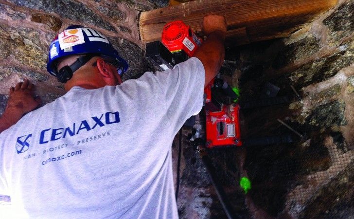 A worker in a gray shirt and blue hard hat uses a red power tool to drill into a stone wall structure.