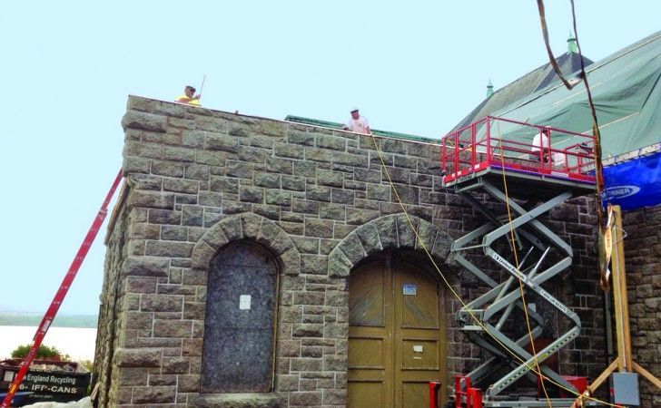 Workers perform stone masonry repairs on a building exterior using a red scissor lift and an extended ladder.
