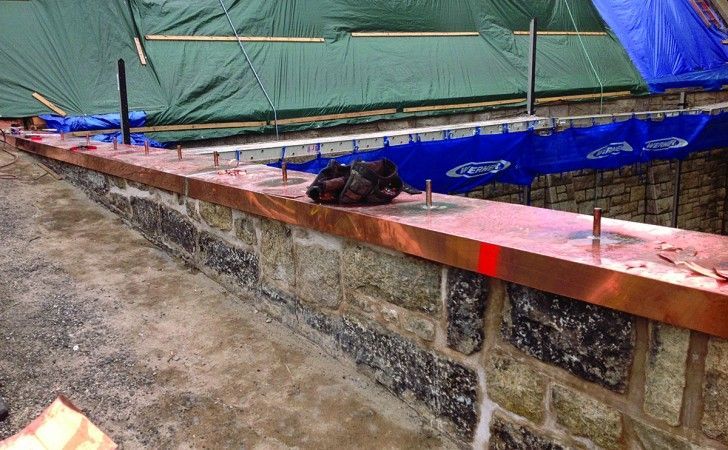 A stone wall under construction with a shiny copper top edge and metal fasteners protruding upwards at regular intervals.