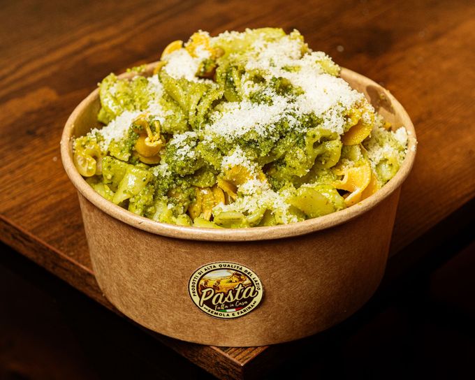 Una ciotola di carta marrone contenente pasta condita con salsa verde e formaggio grattugiato, adagiata su un tavolo di legno.