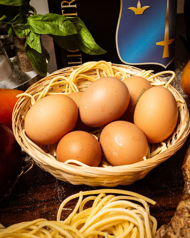 Un cestino di uova marroni con spaghetti crudi su un tavolo scuro, con erbe aromatiche e bottiglie sullo sfondo.