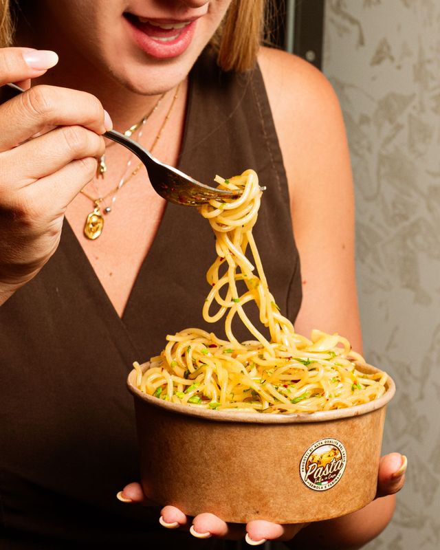Persona che mangia spaghetti da una ciotola con una forchetta, con indosso una maglietta marrone.