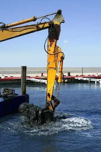 A yellow excavator arm lifting a bucket of dark sediment out of harbor water against a clear blue sky.