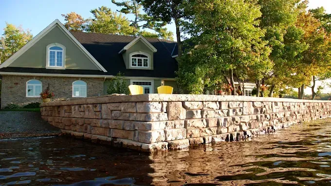 A stone retaining wall along a shoreline in front of a green two-story house surrounded by trees.