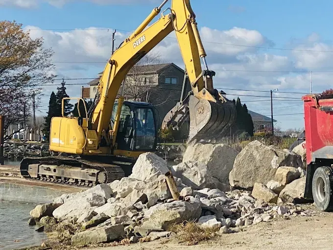 A yellow excavator uses its bucket to move large rocks near a body of water next to a red dump truck.