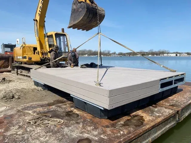 A yellow excavator crane lifts a floating rectangular dock section over a barge at a waterfront construction site.