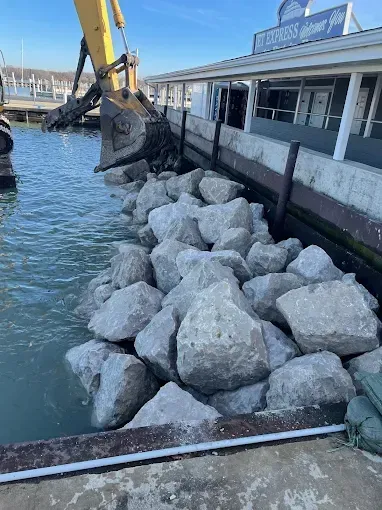 An excavator places large boulders along the waterfront by a restaurant building on a sunny day.