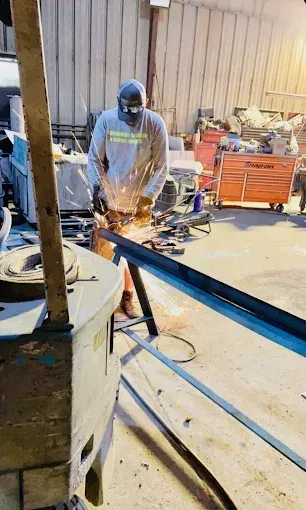 A person in a workshop wears safety gear while using an angle grinder to cut a metal beam, creating a shower of sparks.