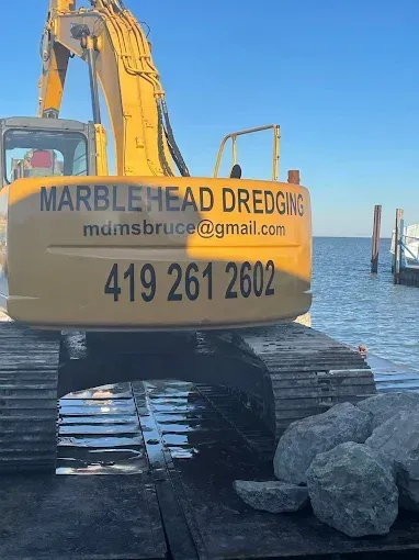A yellow excavator labeled Marblehead Dredging with contact information, parked on a barge next to a body of water.