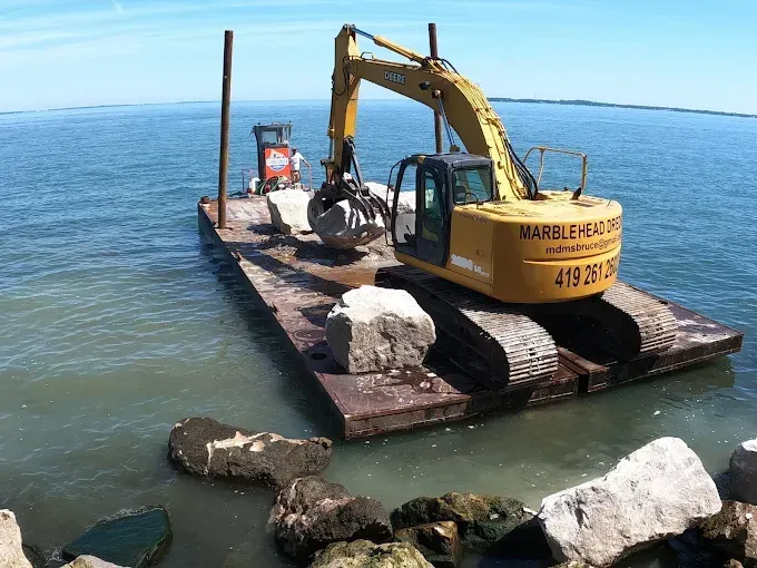 A yellow excavator on a barge in the water, placing large rocks to construct a stone barrier along the shoreline.