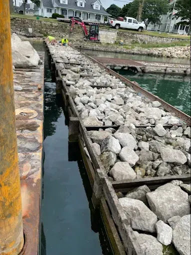 Workers installing a long, rock-filled metal crib structure in a marina, with houses and an excavator visible on shore.