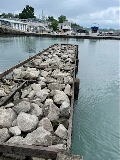 A rectangular, metal-framed dock filled with large, grey rocks projecting into calm harbor water near boats and buildings.