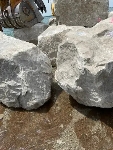 Large, light gray limestone boulders arranged near a waterfront with heavy machinery visible in the background.
