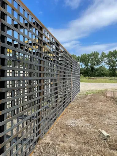 A tall, metal grid fence structure sits outdoors against a blue sky, with a dirt foreground and green trees in the back.