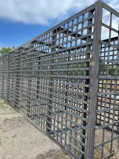 A tall, outdoor metal grid structure or trellis made of dark steel bars, set against a bright, cloudy blue sky.
