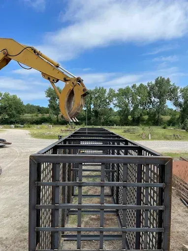 A yellow excavator arm hovers over a long, rectangular metal cage structure outdoors under a blue, cloudy sky.
