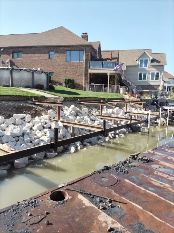 A construction site on a shoreline, featuring metal support beams being installed in front of large rocks and houses.