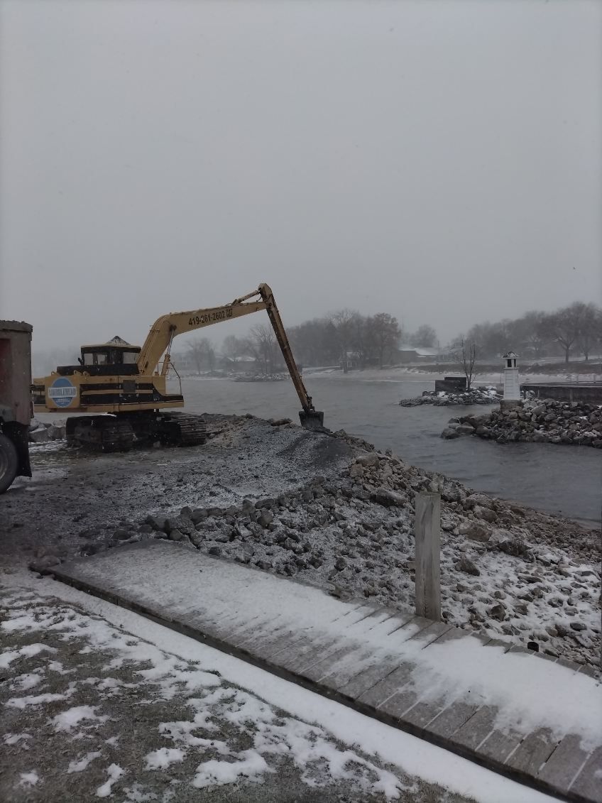 A yellow excavator works on a snowy, rocky shoreline beside a river with a small boat in the background.