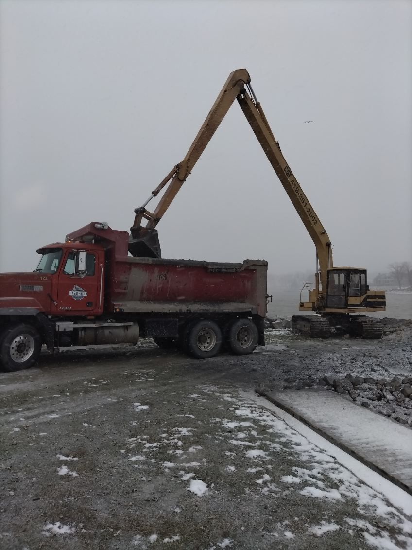 A yellow excavator reaches over a red dump truck, moving rocky material in a snowy, outdoor work site.