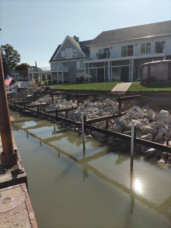 A rocky shoreline with a wooden retaining wall frame along a canal, featuring a large house in the background.