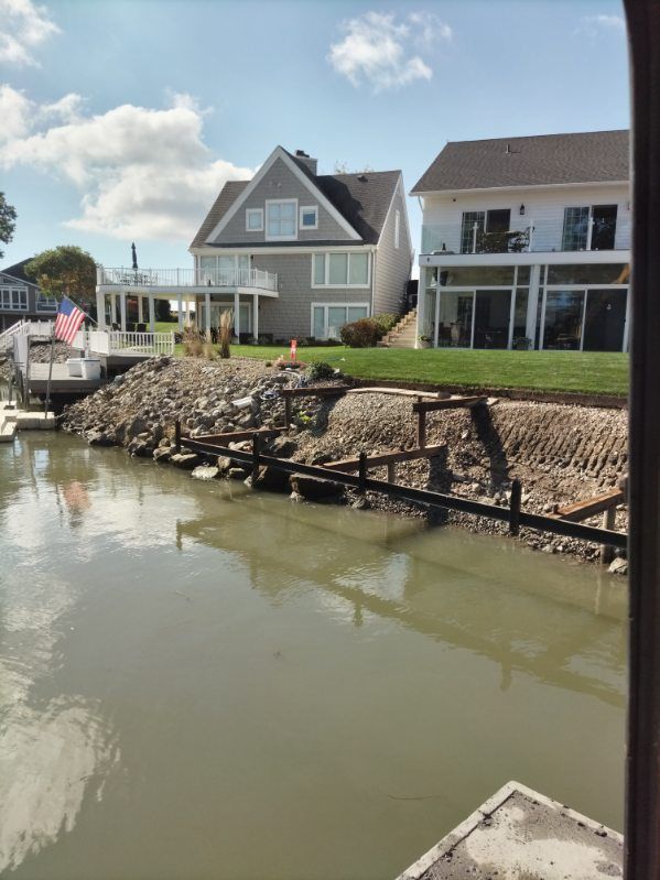 Two waterfront houses with a rocky, uneven shoreline under construction along a canal on a sunny day.