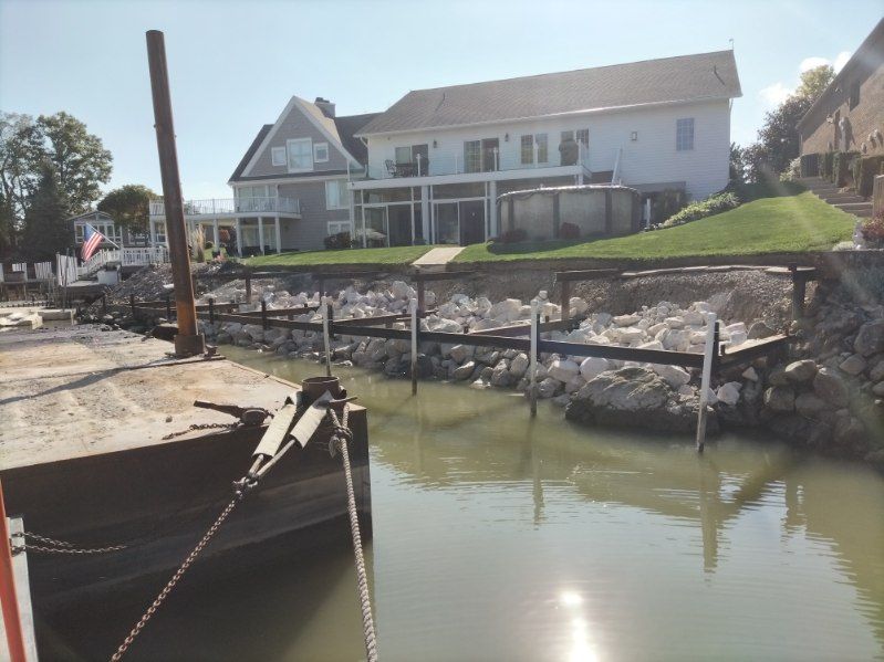 A waterfront property with a new stone retaining wall being constructed along the bank, viewed from a nearby barge.