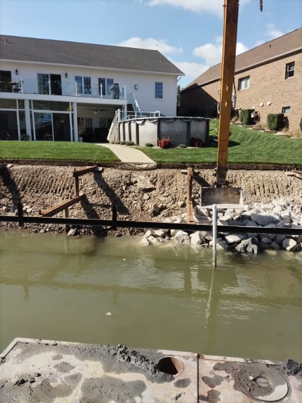 An excavator works on a shoreline embankment near a residential backyard featuring a white house and an above-ground pool.