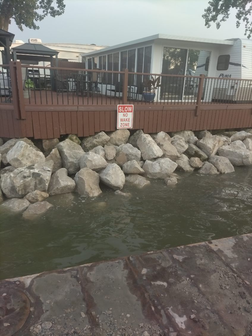 A brown wooden deck overlooking a body of water, with large rocks along the shoreline and a white building in the back.