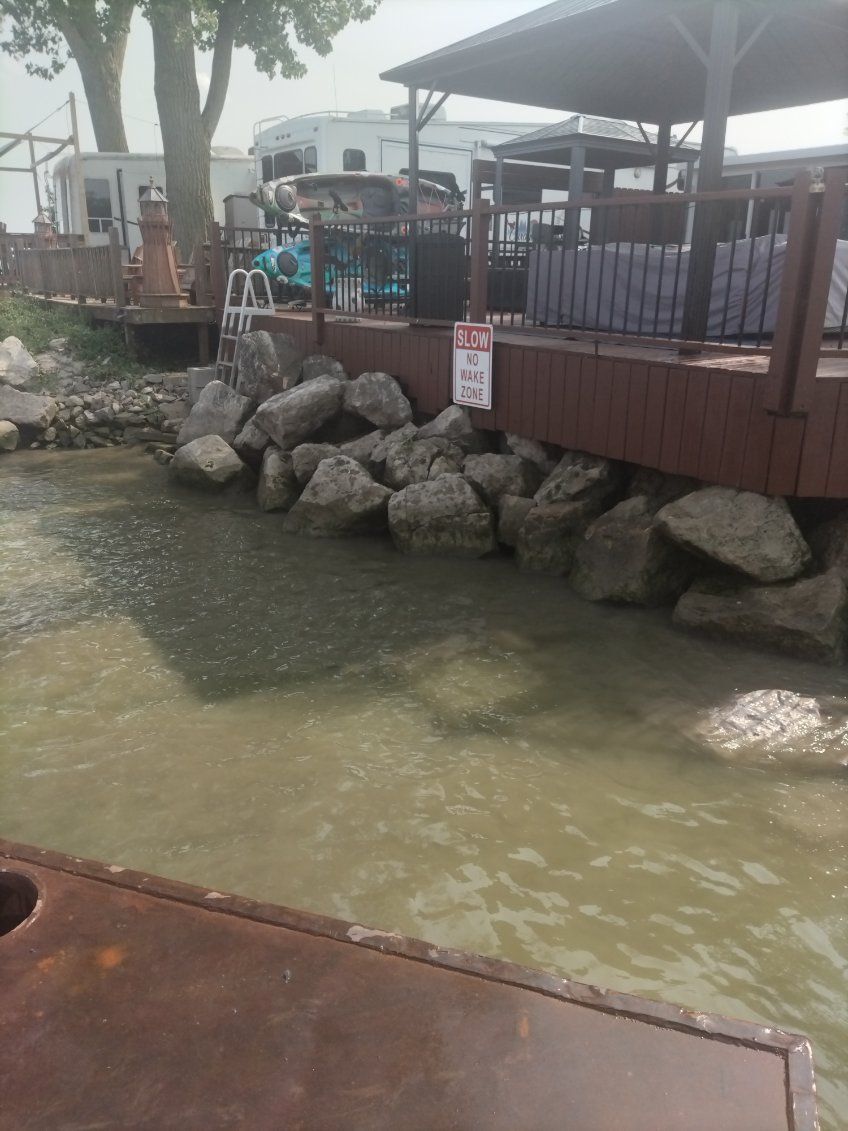 A wooden deck with a gazebo overlooks a rocky shoreline and murky water, with recreational vehicles visible in the back.