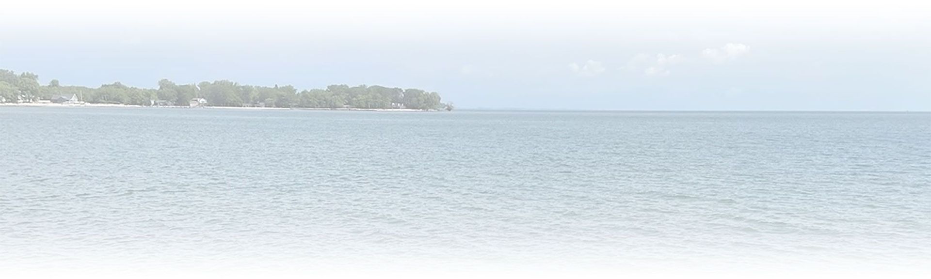 A serene view of a calm blue sea with a distant, forested coastline under a pale, overcast sky.