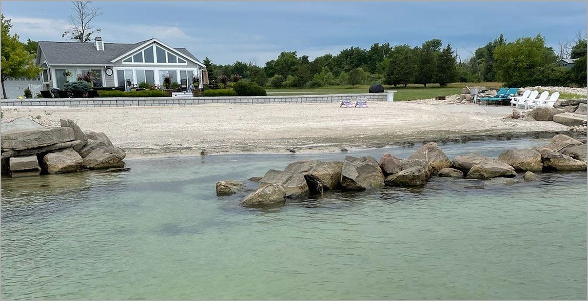 A beach with large rocks in the foreground, a sandy shore, and a white house in the background under a blue sky.