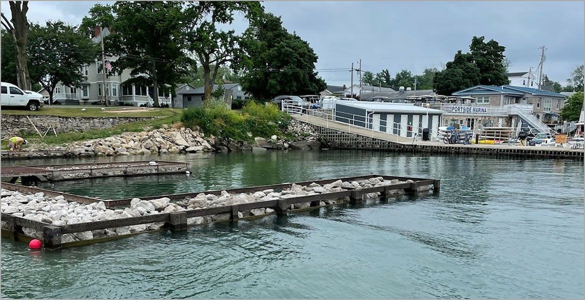 A marina basin with stone-filled wooden crib docks, calm water, and a tree-lined shoreline with buildings in the back.