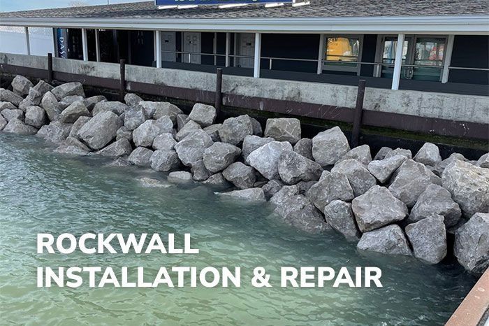 A pile of large gray rocks along a building's foundation by the water, with the text 